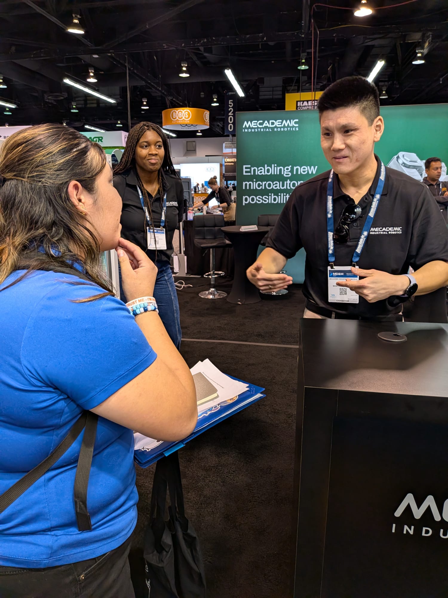 DAYP student in conversation with two Mecademic Industrial Robotics engineers at their MD&M West booth, with green Mecademic backdrop reading 'Enabling new microautomation possibilities'