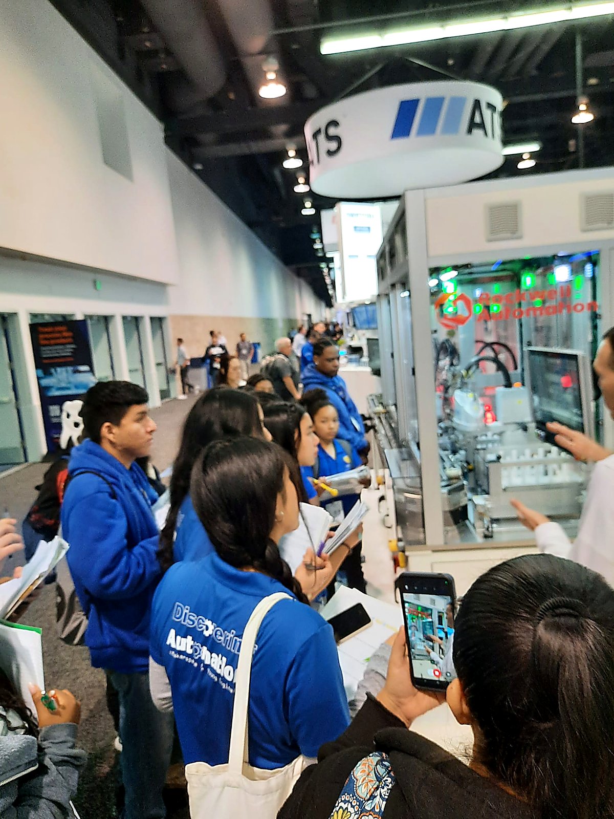 DAYP students observing an Allen-Bradley Rockwell Automation production cell, one student filming with her phone