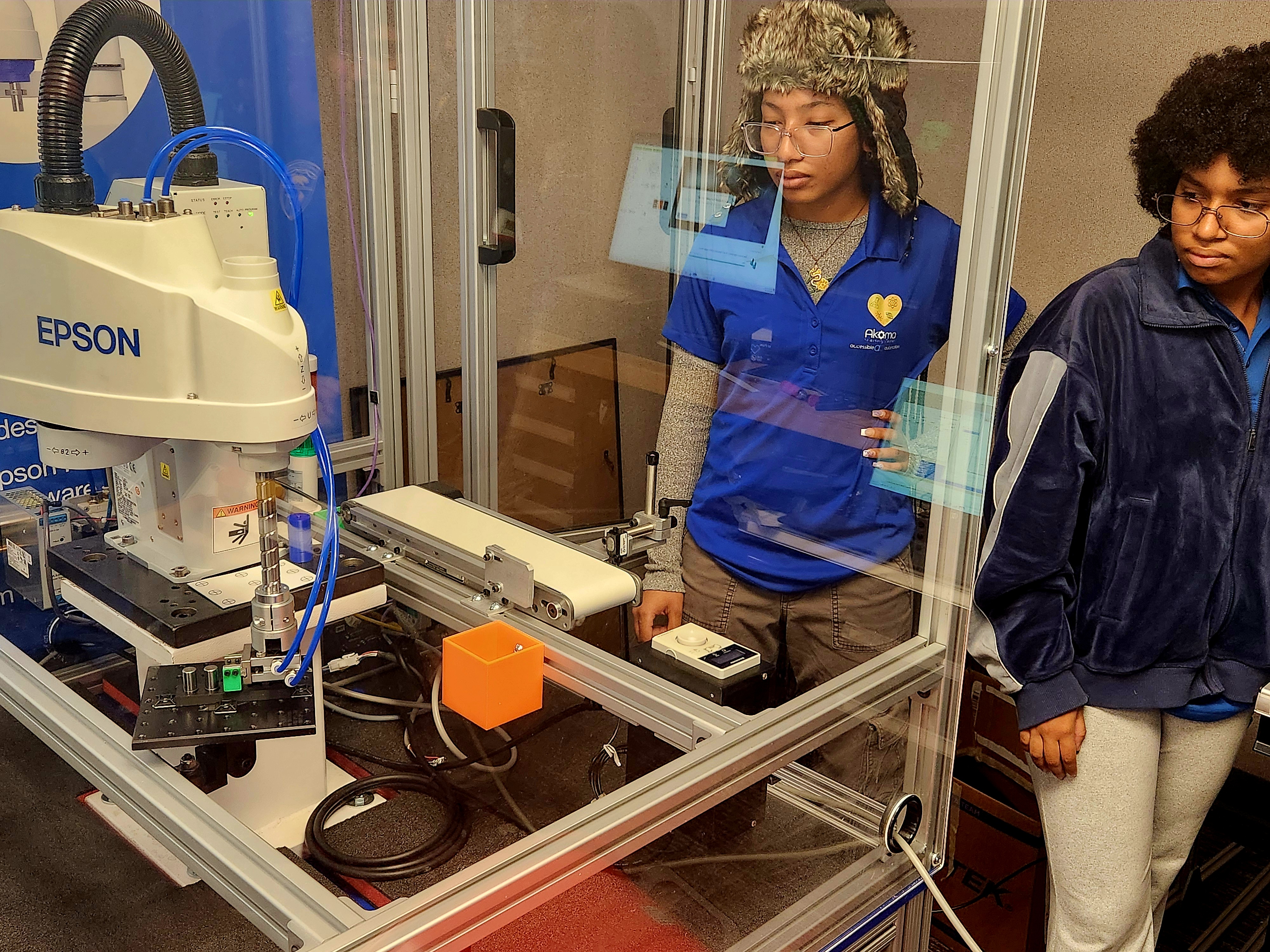 DAYP students observing an Epson SCARA robot running an automated pick-and-place cell with conveyor behind a safety guard