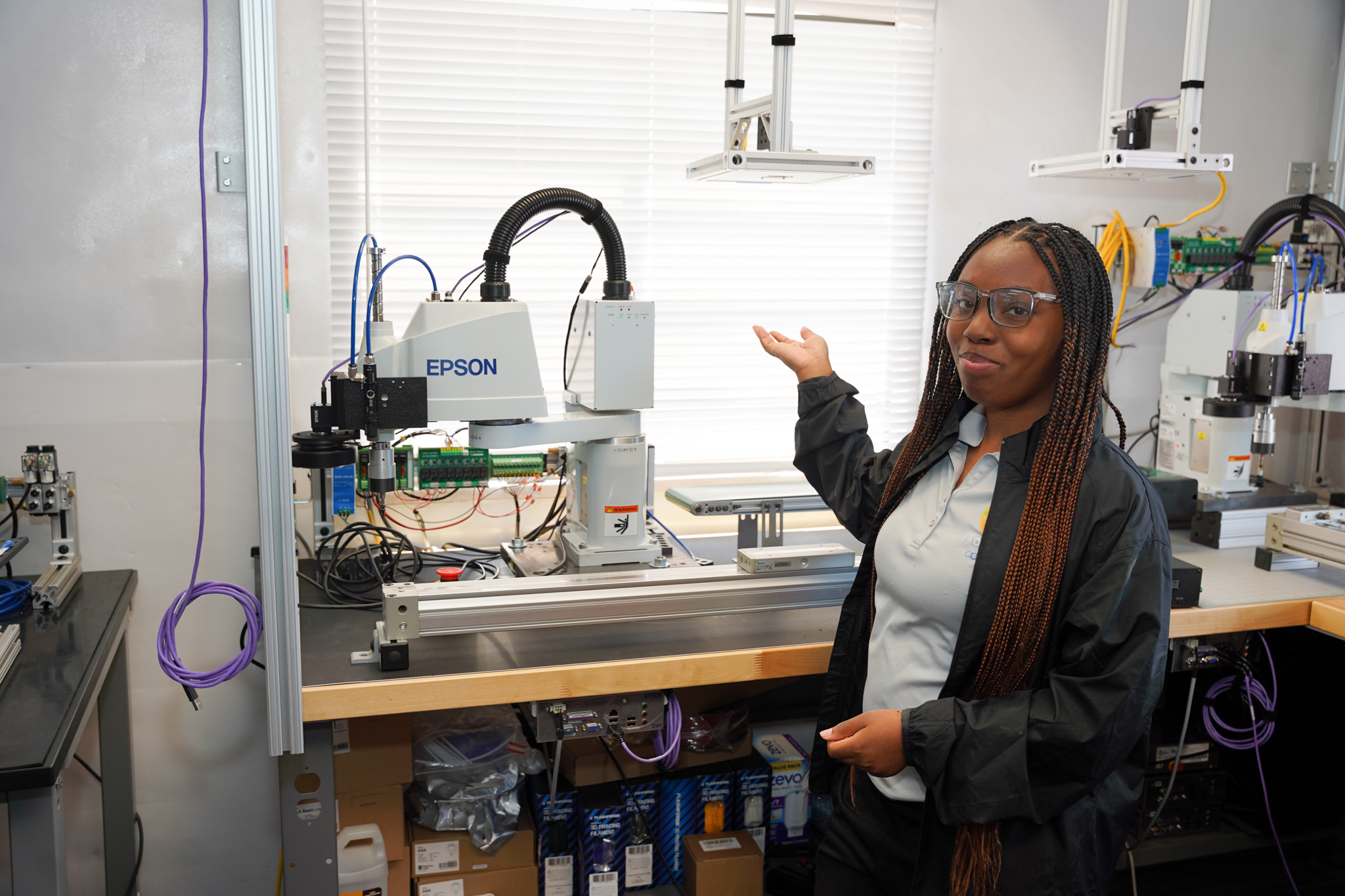 DAYP student in safety glasses standing beside an Epson SCARA robot cell with integrated vision system in the AAI lab, with additional Epson units visible in the background